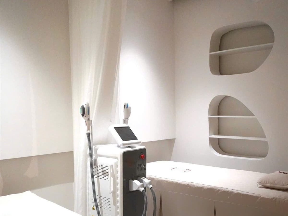 Cabina de estética con dos camillas preparadas, equipo de láser y hornacinas curvas en la pared, todo en tonos claros