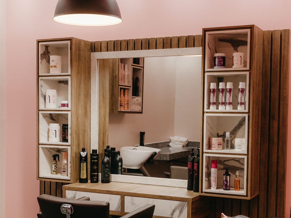 Puesto de peluquería con espejo enmarcado en madera, sillón de corte y estanterías con productos de tratamiento capilar sobre pared rosa
