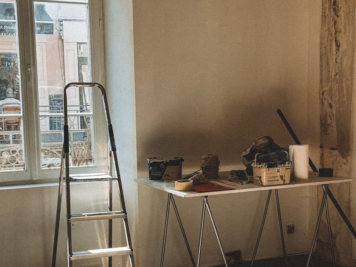 Habitación en obra con escalera de tijera junto a la ventana, mesa de caballetes con botes de pintura, cinta de carrocero y bobina de cuerda, y una pared con el revestimiento antiguo a medio retirar