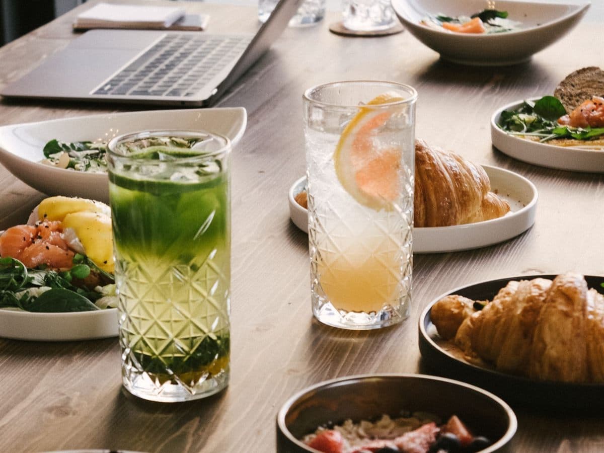 Mesa de brunch servida junto a un ventanal, con platos, bollería y bebidas frías