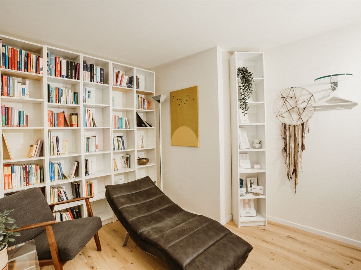 Sala de consulta con una librería completa de suelo a techo, un diván de terapia en tono verde oscuro, butaca de lectura y luz natural sobre suelo de madera clara